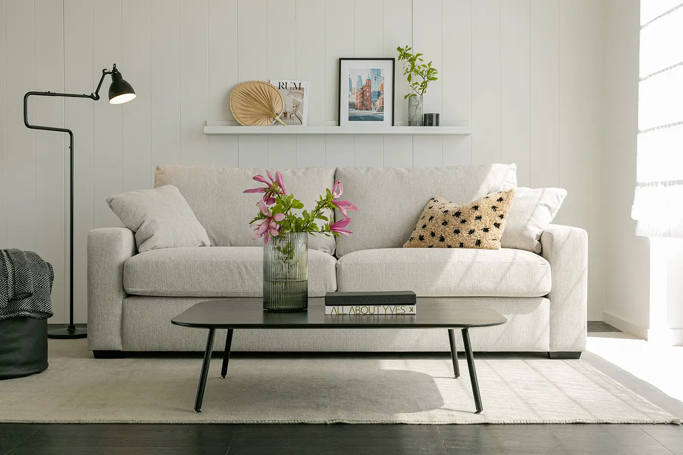 Un salon moderne avec un canapé beige, une table basse, des fleurs dans un vase et des œuvres d'art encadrées sur une étagère blanche. Canape Paris by Atelier Bis