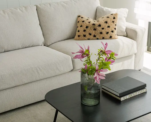 Un canapé beige avec un coussin texturé, une table basse noire, des livres et un vase de fleurs roses dans un salon lumineux. Canape Paris by Atelier Bis