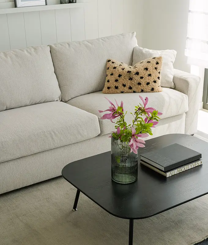 Un canapé beige avec un coussin texturé, une table basse noire, des livres et un vase de fleurs roses dans un salon lumineux. Canape Paris by Atelier Bis