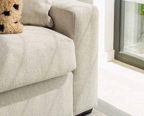 Gros plan sur un canapé gris clair avec un oreiller près d'une fenêtre ensoleillée et d'un rideau blanc transparent. Canape Paris by Atelier Bis