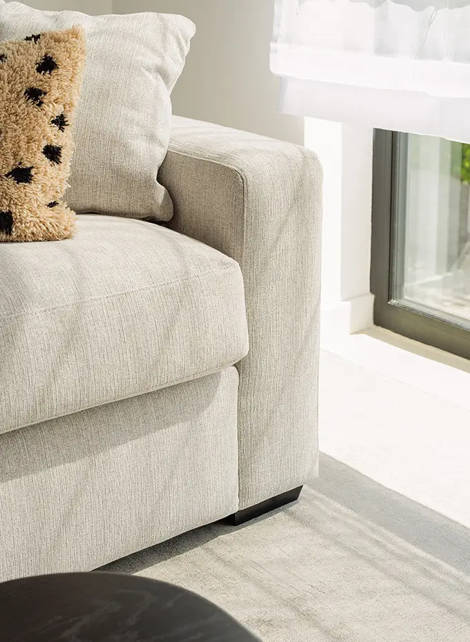 Gros plan sur un canapé gris clair avec un oreiller près d'une fenêtre ensoleillée et d'un rideau blanc transparent. Canape Paris by Atelier Bis