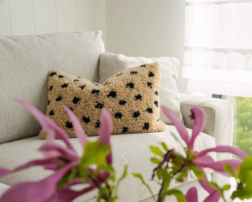 Canapé crème avec un coussin beige à pois noirs près d'une fenêtre ; fleurs roses au premier plan. Canape Paris by Atelier Bis