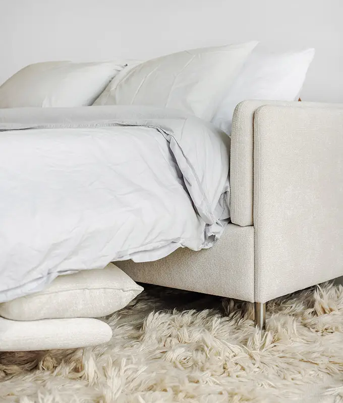 Un lit confortable de couleur claire avec des draps blancs est posé sur un tapis moelleux de couleur crème. Canape Paris by Atelier Bis