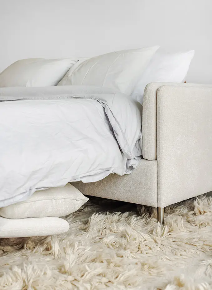 Un lit confortable de couleur claire avec des draps blancs est posé sur un tapis moelleux de couleur crème. Canape Paris by Atelier Bis