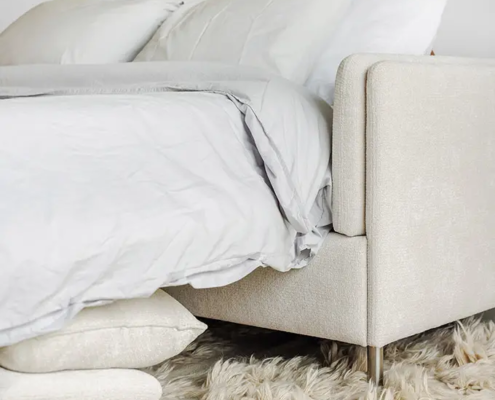 Un lit confortable avec une literie blanche et grise sur un tapis beige moelleux, à côté de deux oreillers posés sur le sol. Canape Paris by Atelier Bis