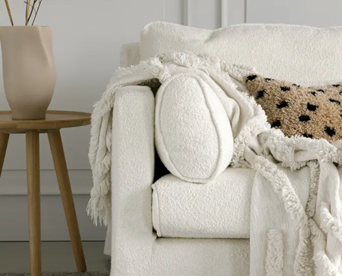 Fauteuil blanc douillet avec un jeté gommé, un oreiller texturé et un vase sur une table d'appoint en bois. Canape Paris by Atelier Bis