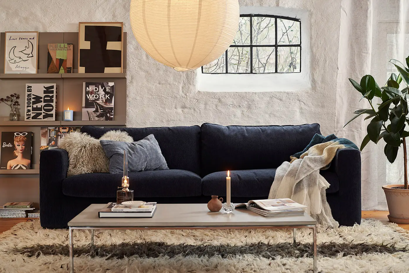 Salon confortable avec un canapé sombre, une table basse, des bougies, une bibliothèque et une grande lampe ronde suspendue. Canape Paris by Atelier Bis