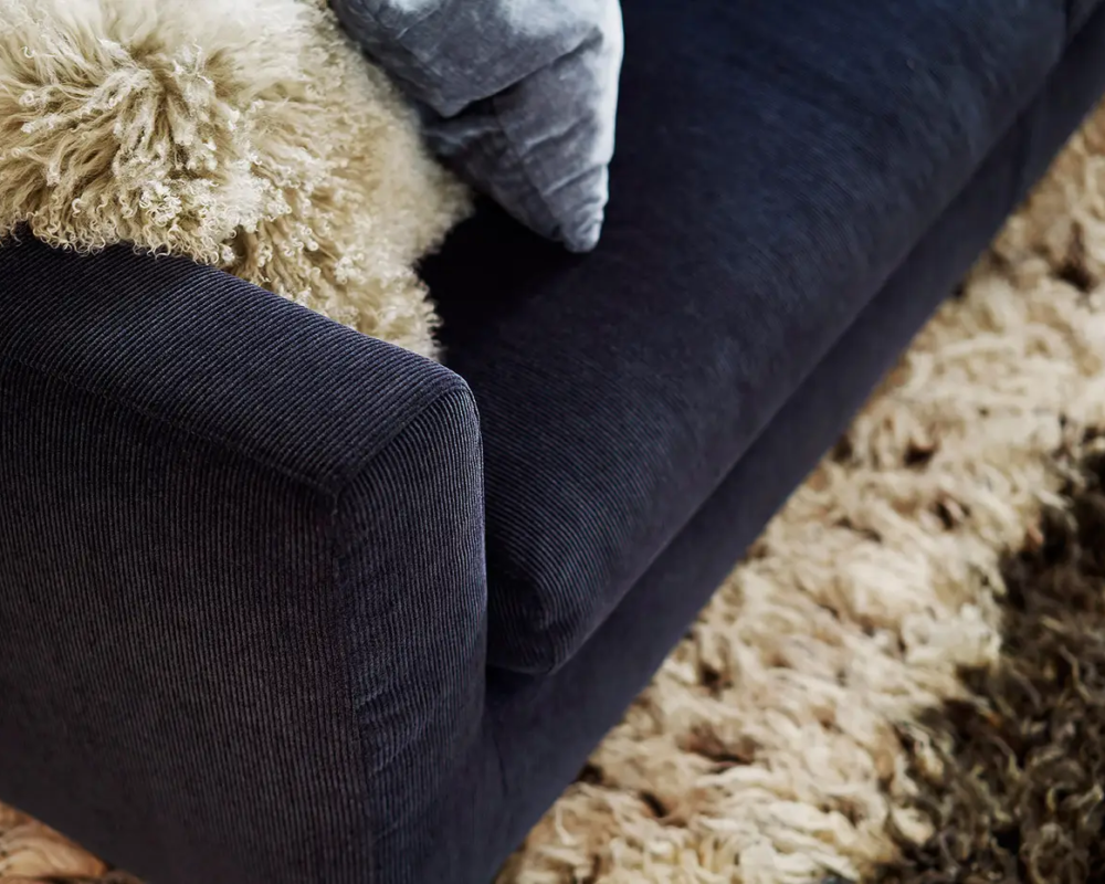 Gros plan sur un canapé bleu foncé avec un jeté et un coussin beige duveteux à côté d'un tapis beige hirsute. Canape Paris by Atelier Bis