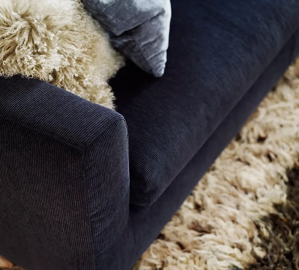 Gros plan sur un canapé bleu foncé avec un jeté et un coussin beige duveteux à côté d'un tapis beige hirsute. Canape Paris by Atelier Bis