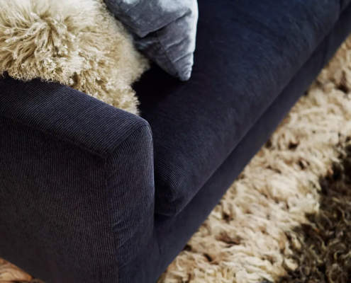 Gros plan sur un canapé bleu foncé avec un jeté et un coussin beige duveteux à côté d'un tapis beige hirsute. Canape Paris by Atelier Bis