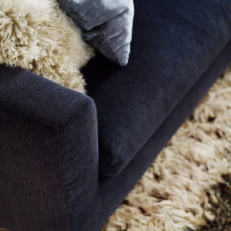 Gros plan sur un canapé bleu foncé avec un jeté et un coussin beige duveteux à côté d'un tapis beige hirsute. Canape Paris by Atelier Bis