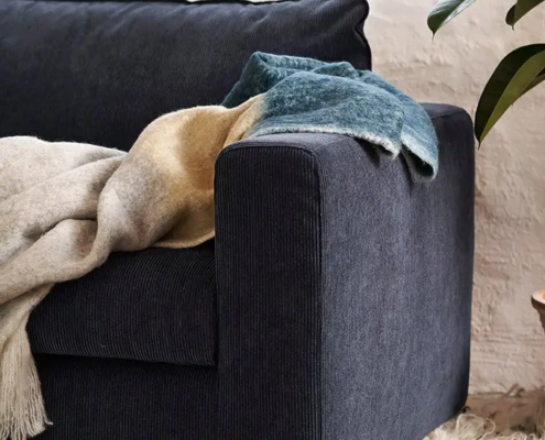 Canapé bleu foncé en velours côtelé surmonté de couvertures douillettes, à côté d'une plante et d'un tapis texturé. Canape Paris by Atelier Bis
