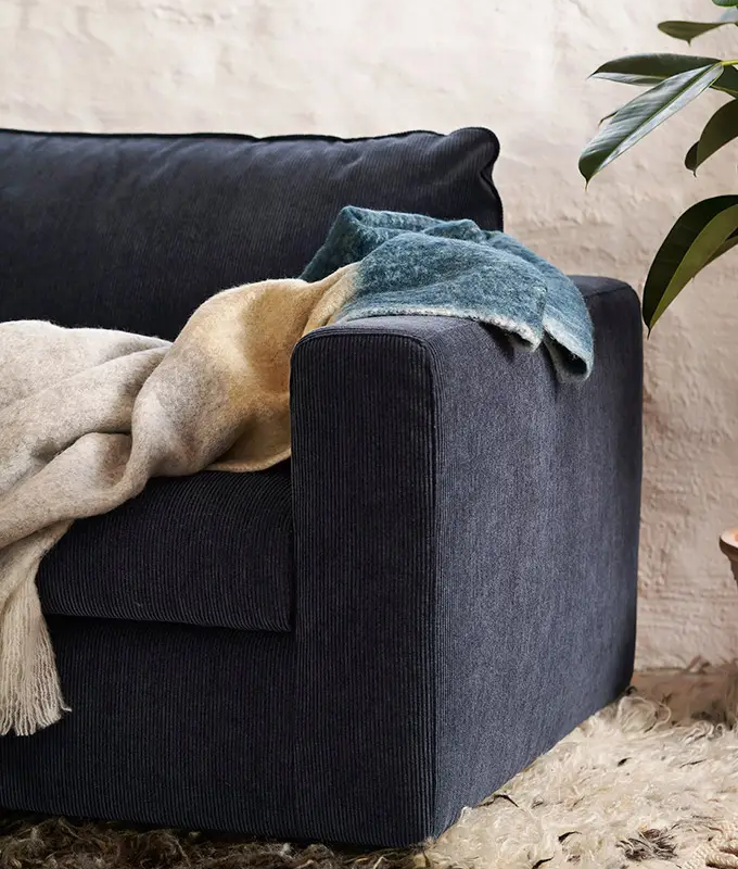 Canapé bleu foncé en velours côtelé surmonté de couvertures douillettes, à côté d'une plante et d'un tapis texturé. Canape Paris by Atelier Bis