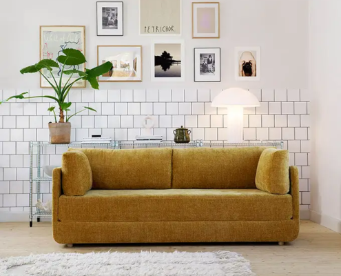 Canapé jaune moutarde dans un salon moderne avec art mural, plante d'intérieur, lampe et tapis blanc sur sol en bois. Canape Paris by Atelier Bis