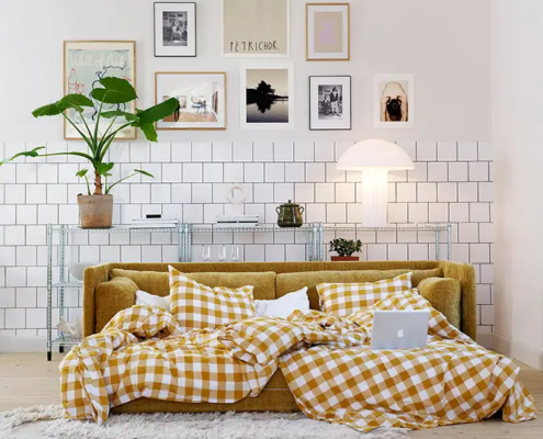Un canapé-lit confortable avec une literie à carreaux jaunes, un ordinateur portable et des coussins dans un salon moderne et élégant. Canape Paris by Atelier Bis