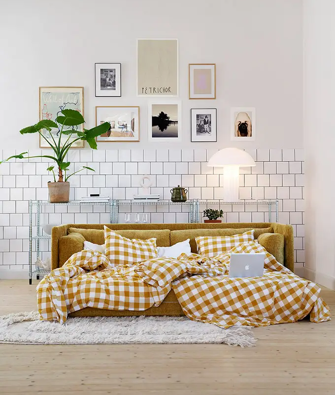 Un canapé-lit confortable avec une literie à carreaux jaunes, un ordinateur portable et des coussins dans un salon moderne et élégant. Canape Paris by Atelier Bis