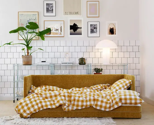 Un canapé jaune moutarde avec une couverture vichy jaune dans un salon moderne et lumineux avec des œuvres d'art murales et des plantes. Canape Paris by Atelier Bis
