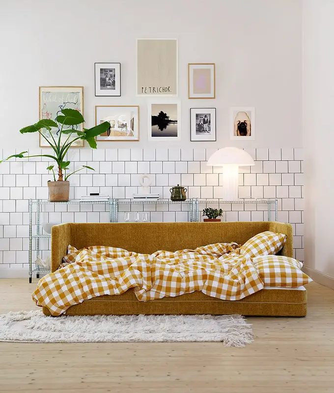 Un canapé jaune moutarde avec une couverture vichy jaune dans un salon moderne et lumineux avec des œuvres d'art murales et des plantes. Canape Paris by Atelier Bis