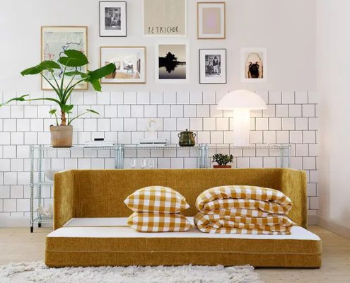 Un salon moderne avec un canapé-lit jaune moutarde, des oreillers à carreaux et un mur d'art au-dessus. Canape Paris by Atelier Bis