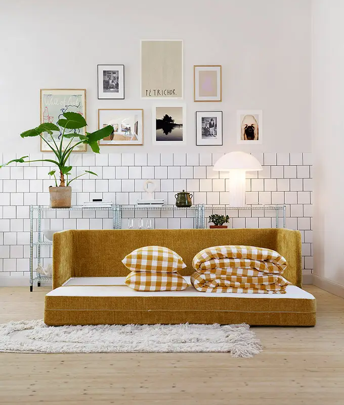Un salon moderne avec un canapé-lit jaune moutarde, des oreillers à carreaux et un mur d'art au-dessus. Canape Paris by Atelier Bis