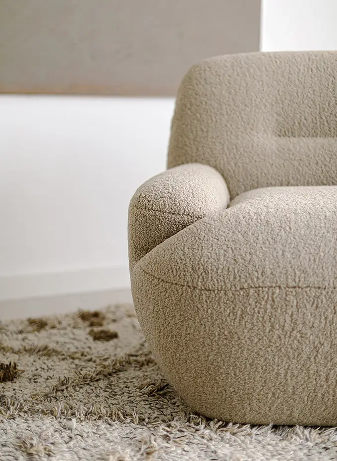 Gros plan sur un fauteuil beige et texturé posé sur un tapis shaggy dans une pièce minimaliste. Canape Paris by Atelier Bis