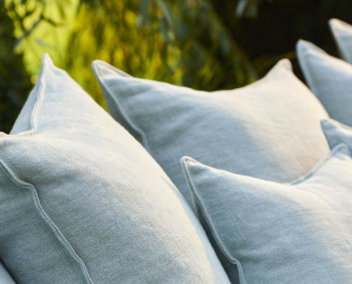 Gros plan sur plusieurs coussins en lin gris clair disposés à l'extérieur avec de la verdure en arrière-plan. Canape Paris by Atelier Bis