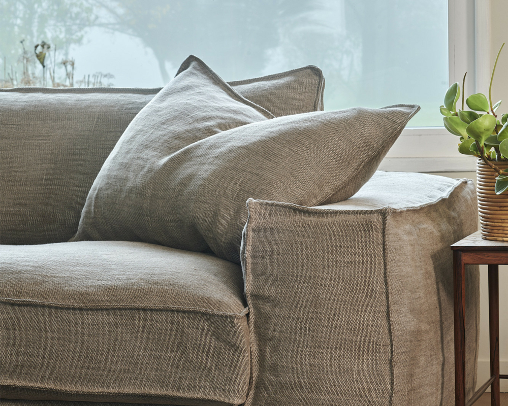 Un canapé en lin beige avec de gros coussins à côté d'une petite table en bois et une plante verte près d'une fenêtre. Canape Paris by Atelier Bis