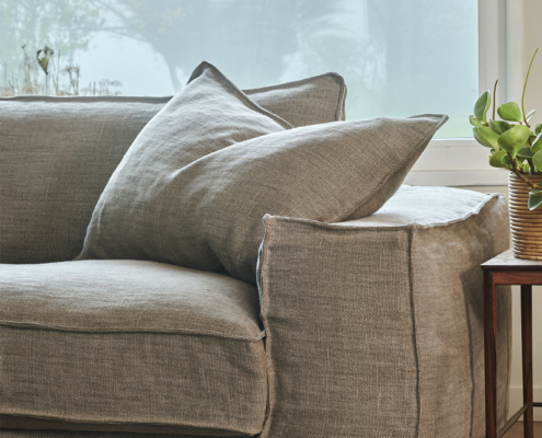 Un canapé en lin beige avec de gros coussins à côté d'une petite table en bois et une plante verte près d'une fenêtre. Canape Paris by Atelier Bis
