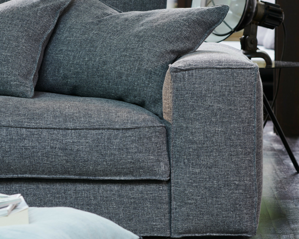 Gros plan sur un canapé gris moderne avec de gros coussins, un lampadaire et un coussin bleu clair posé sur le sol à proximité. Canape Paris by Atelier Bis