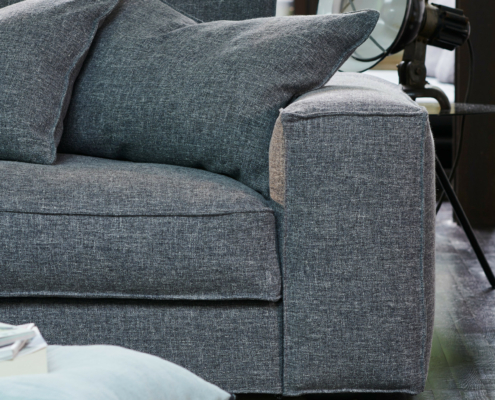 Gros plan sur un canapé gris moderne avec de gros coussins, un lampadaire et un coussin bleu clair posé sur le sol à proximité. Canape Paris by Atelier Bis