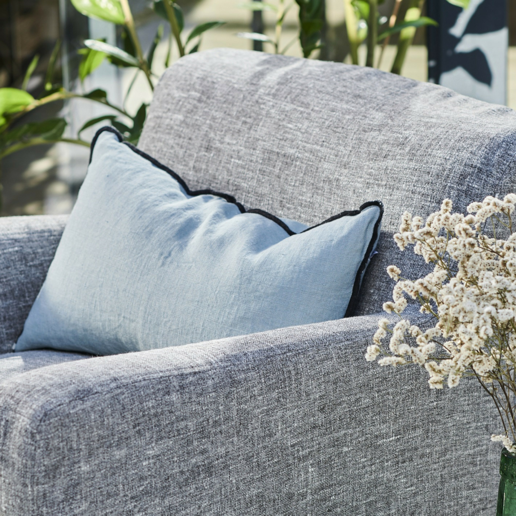 Un fauteuil gris avec un coussin bleu clair et un vase de fleurs blanches dans une pièce ensoleillée avec des plantes. Canape Paris by Atelier Bis