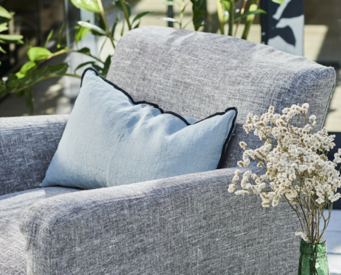 Un fauteuil gris avec un coussin bleu clair et un vase de fleurs blanches dans une pièce ensoleillée avec des plantes. Canape Paris by Atelier Bis