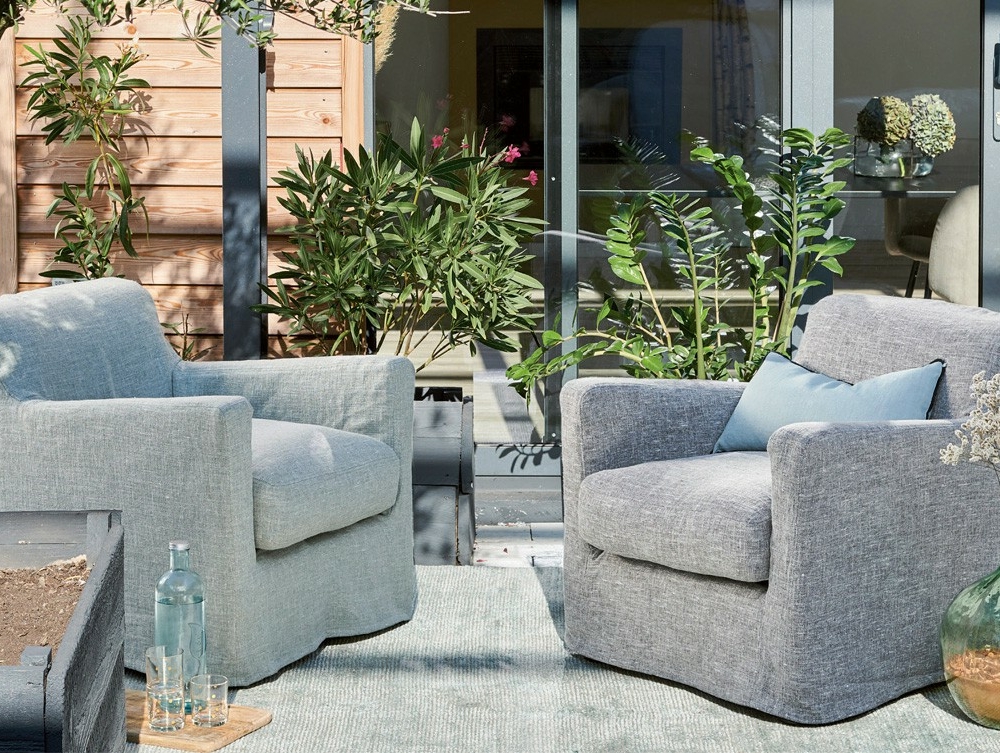 Deux fauteuils gris et un coussin bleu sur un patio, entouré de plantes et de portes vitrées. Canape Paris by Atelier Bis