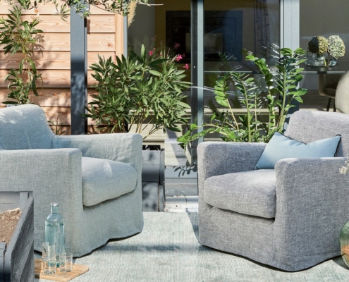 Deux fauteuils gris et un coussin bleu sur un patio, entouré de plantes et de portes vitrées. Canape Paris by Atelier Bis