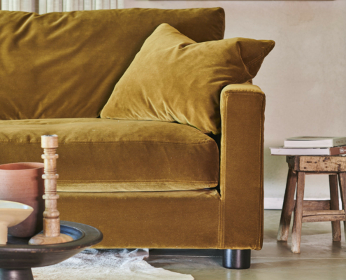 Canapé en velours jaune moutarde avec coussin, à côté d'un tabouret rustique et de livres sur des étagères dans un salon confortable. Canape Paris by Atelier Bis