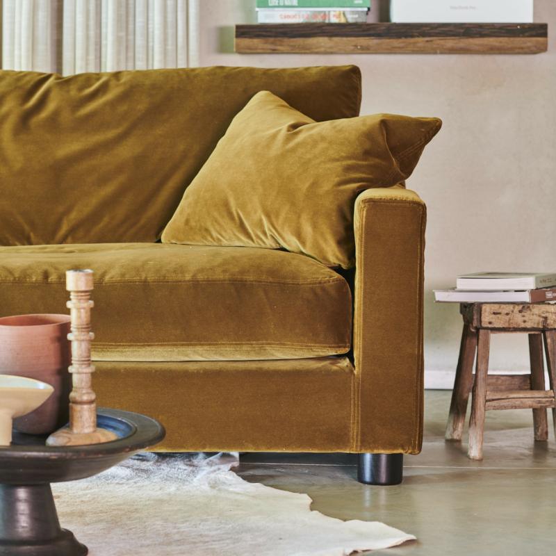 Canapé en velours jaune moutarde avec coussin, à côté d'un tabouret rustique et de livres sur des étagères dans un salon confortable. Canape Paris by Atelier Bis
