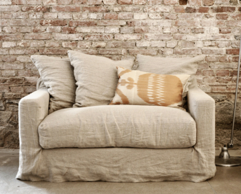 Fauteuil beige avec trois coussins contre un mur rustique en briques et un lampadaire à côté. Canape Paris by Atelier Bis