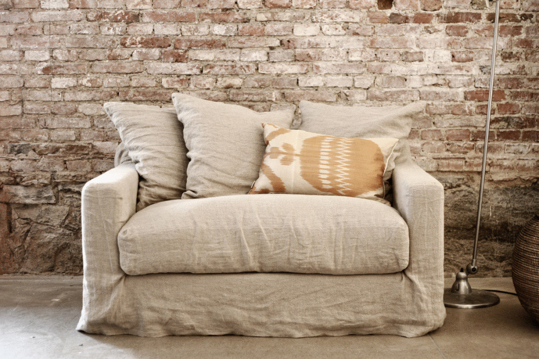 Fauteuil beige avec trois coussins contre un mur rustique en briques et un lampadaire à côté. Canape Paris by Atelier Bis