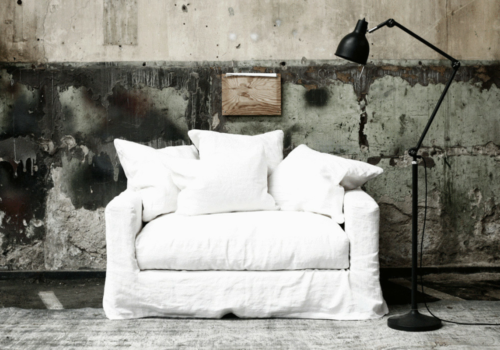 Un canapé blanc avec des coussins et un lampadaire noir contre un mur industriel en béton vieilli. Canape Paris by Atelier Bis