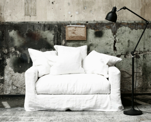 Un canapé blanc avec des coussins et un lampadaire noir contre un mur industriel en béton vieilli. Canape Paris by Atelier Bis