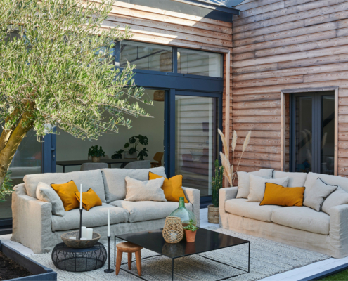 Un patio extérieur confortable avec des canapés beiges, des coussins jaunes, une table basse noire et un grand arbre en pot à proximité. Canape Paris by Atelier Bis