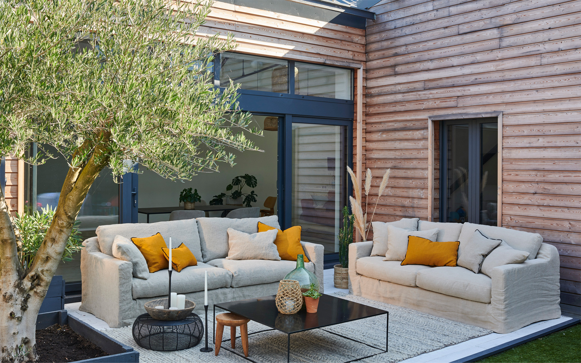 Un patio extérieur confortable avec des canapés beiges, des coussins jaunes, une table basse noire et un grand arbre en pot à proximité. Canape Paris by Atelier Bis