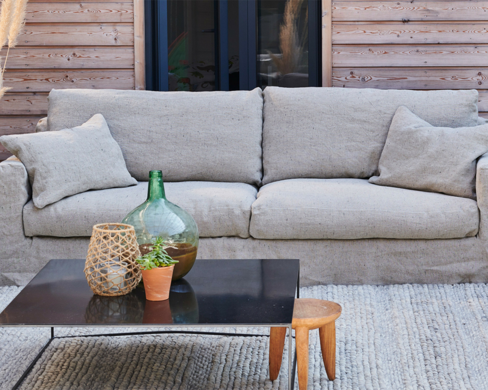 Canapé beige avec coussins sur un patio, table basse noire avec plantes, et tabouret en bois devant. Canape Paris by Atelier Bis
