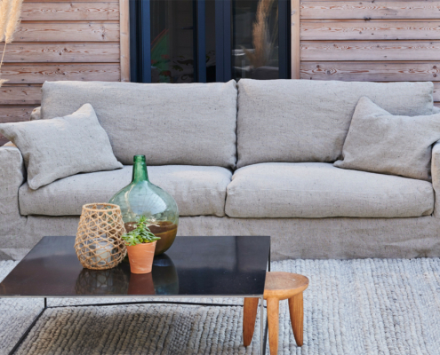 Canapé beige avec coussins sur un patio, table basse noire avec plantes, et tabouret en bois devant. Canape Paris by Atelier Bis