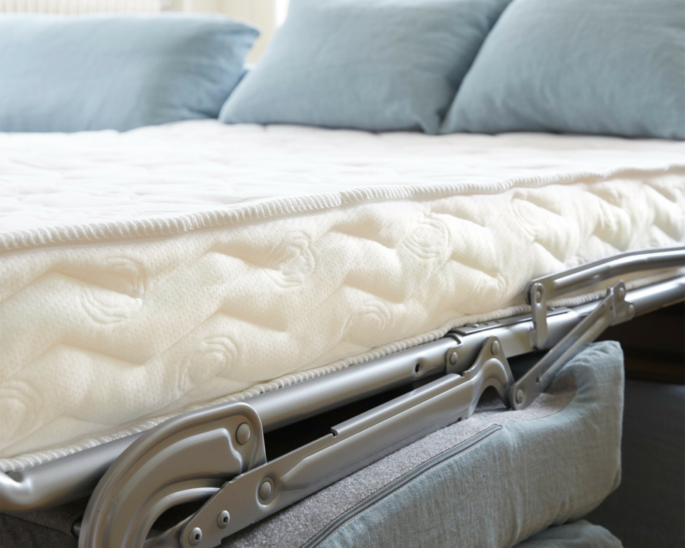 Gros plan sur un lit d'invité pliant avec des oreillers bleus et un matelas blanc dans une pièce lumineuse. Canape Paris by Atelier Bis