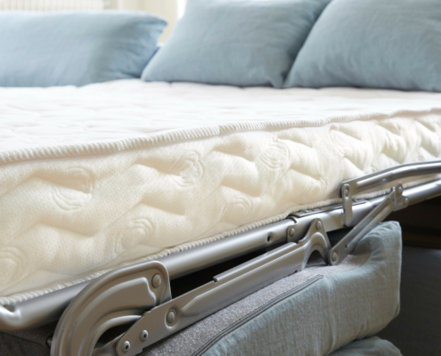 Gros plan sur un lit d'invité pliant avec des oreillers bleus et un matelas blanc dans une pièce lumineuse. Canape Paris by Atelier Bis