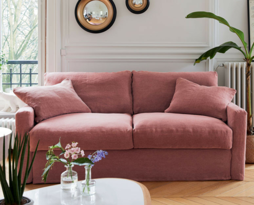 Un canapé rose avec deux coussins dans un salon lumineux avec des plantes, des miroirs et une grande fenêtre. Canape Paris by Atelier Bis