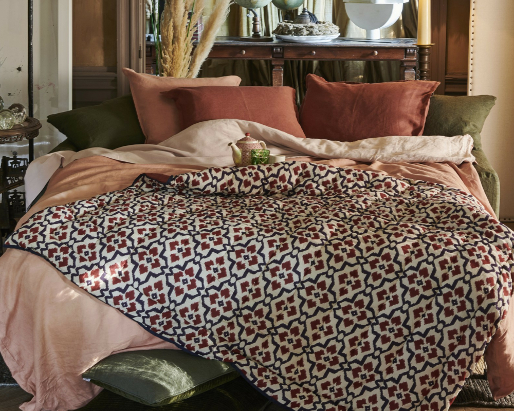 Lit douillet avec des oreillers roses, verts et rouille, et une couverture à motifs rouges et noirs dans une pièce chaudement éclairée. Canape Paris by Atelier Bis