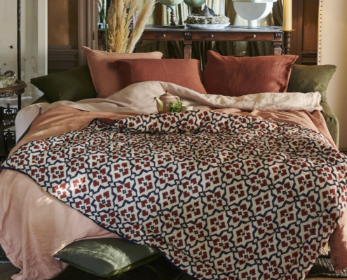 Lit douillet avec des oreillers roses, verts et rouille, et une couverture à motifs rouges et noirs dans une pièce chaudement éclairée. Canape Paris by Atelier Bis