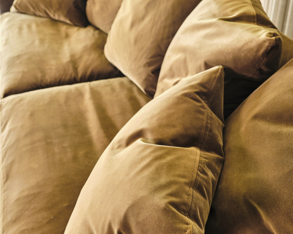 Un canapé en velours marron avec des coussins en peluche assortis, placé près d'une fenêtre avec de la verdure à l'extérieur. Canape Paris by Atelier Bis